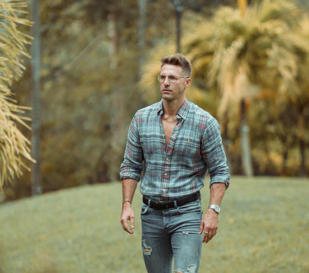 Thomas Beattie walking outdoors while wearing a flannel shirt and jeans. A green landscape can be seen in the background.