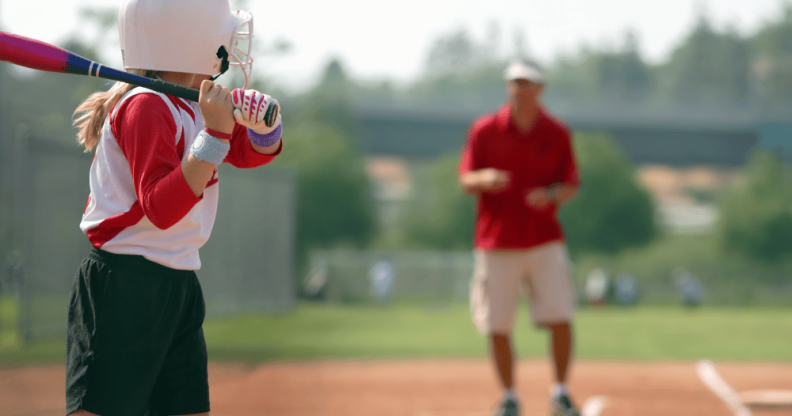 Stock image of a young girl holding a bat while a man coaches her in the background