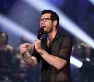 Billy Eichner wears a dark outfit as he gestures towards himself as he speaks at the MTV VMAs