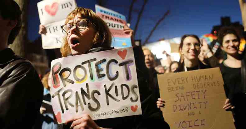 A person in gather amid a crowd in a protest holding a sign that reads 'Protect trans kids'