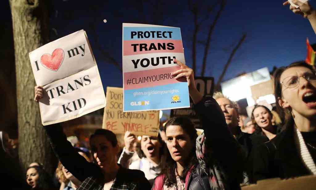 A person is in a gathering amid a crowd in a protest holding a sign that reads 'Protect trans kids'