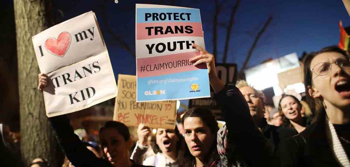 A person is in a gathering amid a crowd in a protest holding a sign that reads 'Protect trans kids'