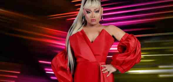 In this photograph, Drag Race Philippines host Jiggly Caliente wears a red sleeveless dress with matching red fabric pieces on her forearms. She has long blonde and brown hair styled in a ponytail and is posed with one hand on her hip. She is standing in front of a dark background with lines of pink, red and yellow