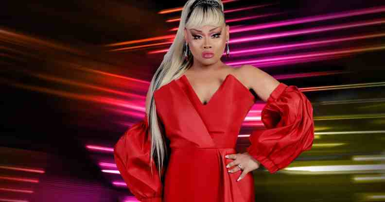 In this photograph, Drag Race Philippines host Jiggly Caliente wears a red sleeveless dress with matching red fabric pieces on her forearms. She has long blonde and brown hair styled in a ponytail and is posed with one hand on her hip. She is standing in front of a dark background with lines of pink, red and yellow