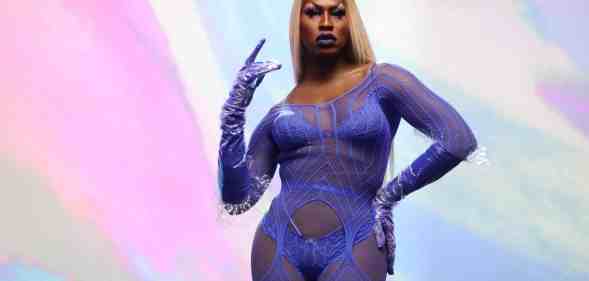 Shea Coulee stands in front of a multi-coloured background wearing a purple lace lingerie jumpsuit.