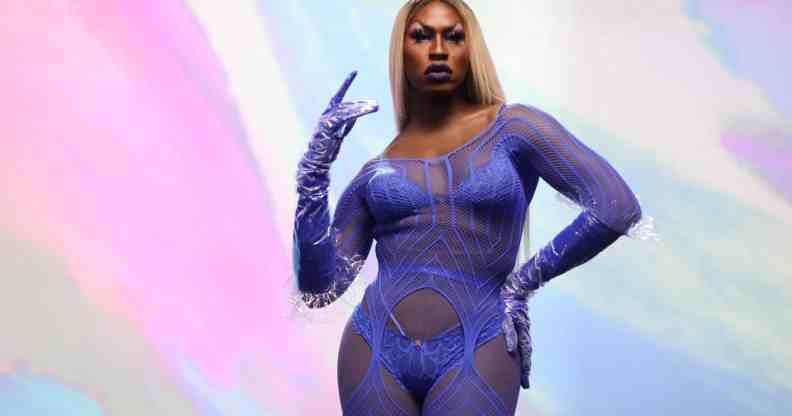 Shea Coulee stands in front of a multi-coloured background wearing a purple lace lingerie jumpsuit.