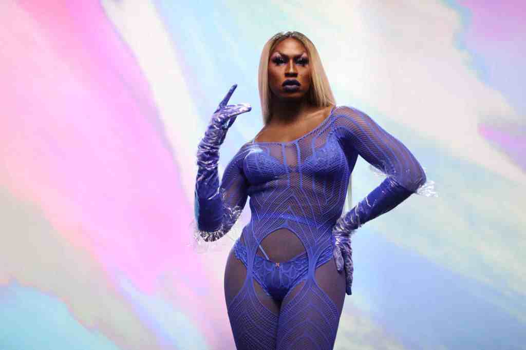 Shea Coulee stands in front of a multi-coloured background wearing a purple lace lingerie jumpsuit.