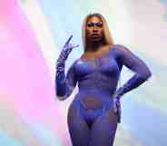 Shea Coulee stands in front of a multi-coloured background wearing a purple lace lingerie jumpsuit.