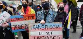 Several people join a protest outside the BBC. One person holds a sign that reads 'Transphobia is not unbiased' that is coloured with blue, pink and white stripes