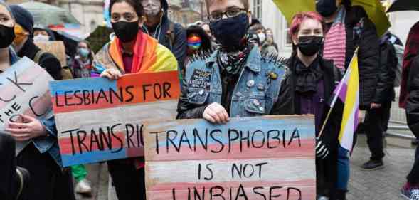 Several people join a protest outside the BBC. One person holds a sign that reads 'Transphobia is not unbiased' that is coloured with blue, pink and white stripes