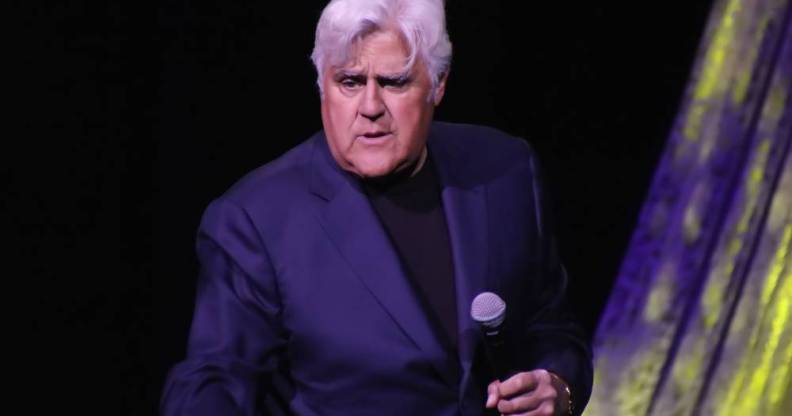 Jay Leno holds a microphone in his hand as he wears a black shirt and dark blue suit jacket as he stands on stage