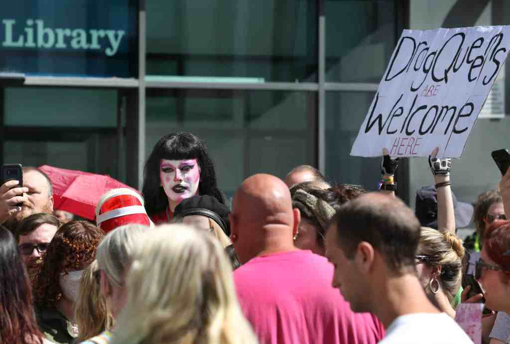 A crowd protesting against the exclusion of drag queens. One person holds a sign that says, "Drag queens welcome here"