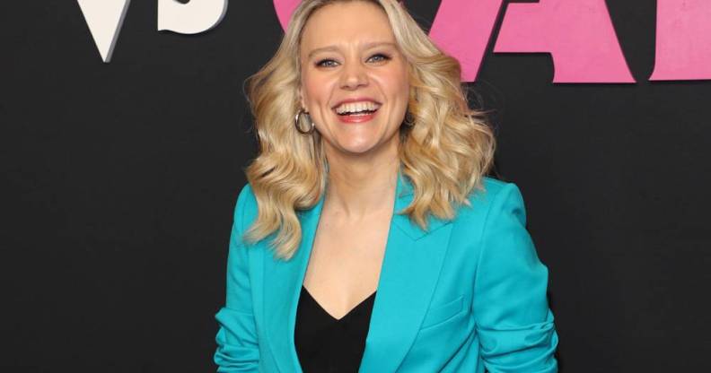 Kate McKinnon smiles at the camera while wearing a black top and bright blue blazer on top. She is standing in front of a black background with pink and white writing on it