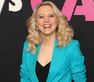 Kate McKinnon smiles at the camera while wearing a black top and bright blue blazer on top. She is standing in front of a black background with pink and white writing on it