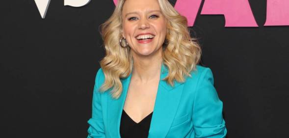 Kate McKinnon smiles at the camera while wearing a black top and bright blue blazer on top. She is standing in front of a black background with pink and white writing on it