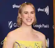 Tommy Dorfman stands in the press section of the GLAAD Media Awards wearing a yellow dress.