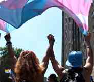 In this photograph, two trans people hold hands while marching under a trans pride flag