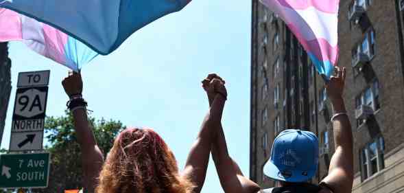 In this photograph, two trans people hold hands while marching under a trans pride flag