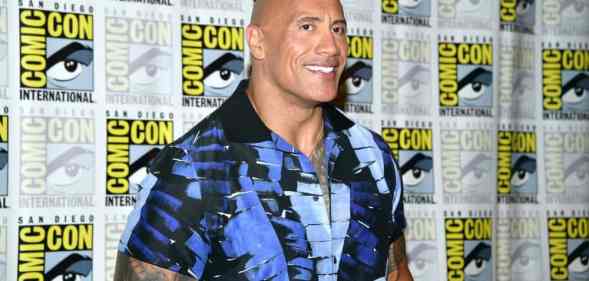 Dwayne 'The Rock' Johnson smiles for the camera as he wears a blue and black patterned top and stands in front of a patterned background with the SD Comic Con logo on it