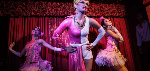 Three drag queens on a stage performing wearing pink dresses.