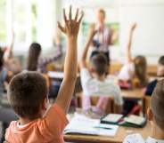 A stock photo of children with their hands raised in a classroom, facing a teacher and a whiteboard
