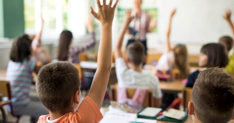 A stock photo of children with their hands raised in a classroom, facing a teacher and a whiteboard