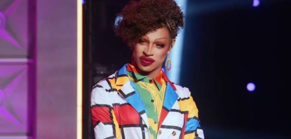 Drag Race star Yvie Oddly wears a white, blue, red and yellow geometric patterned outler layer with another colourful layer of clothing below. Yvie's hair is styled in an updo
