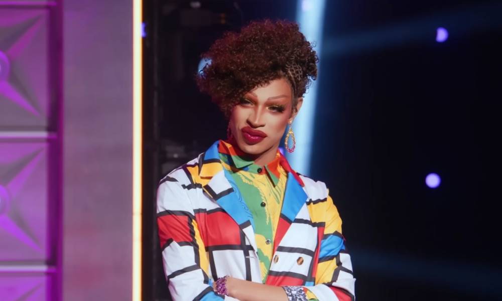 Drag Race star Yvie Oddly wears a white, blue, red and yellow geometric patterned outler layer with another colourful layer of clothing below. Yvie's hair is styled in an updo
