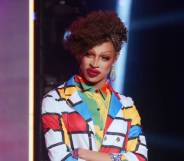Drag Race star Yvie Oddly wears a white, blue, red and yellow geometric patterned outler layer with another colourful layer of clothing below. Yvie's hair is styled in an updo