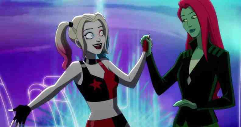 A still taken from the Harley Quinn animated series on HBO Max. In this image, Harley Quinn wears a black and red patterned outfit as she holds the hand of Poison Ivy, who is wearing dark clothing and staring at Harley