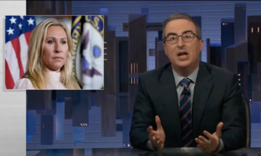 John Oliver wears a blue shirt, striped tie and dark suit jacket on the set of HBO Max's Last Week Tonight. There is an image of Georgia congresswoman Marjorie Taylor Greene in the corner. Greene is wearing a white top as she stands in front of a US flag