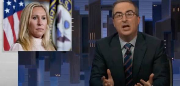 John Oliver wears a blue shirt, striped tie and dark suit jacket on the set of HBO Max's Last Week Tonight. There is an image of Georgia congresswoman Marjorie Taylor Greene in the corner. Greene is wearing a white top as she stands in front of a US flag