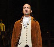 Lin-Manuel Miranda wears a white shirt, cream coloured waist jacket and brown coat as he performs in Hamilton