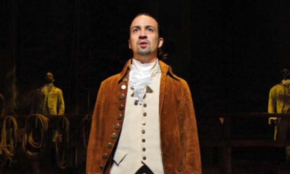 Lin-Manuel Miranda wears a white shirt, cream coloured waist jacket and brown coat as he performs in Hamilton