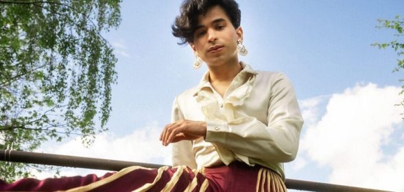 Shiva Raichandani in a white top and red and gold trousers leaning against a fence.
