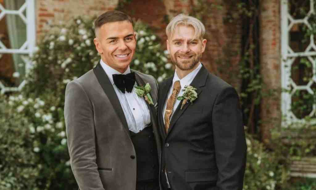 A screenshot of Thomas and Adrian dressed in their wedding outfits from Married at First Sight UK. Thomas' grey suit looks to be made from velvet and he's wearing a formal white shirt and black bowtie and Adrian is dressed in a more conventional dark-looking suit with patterned tie.
