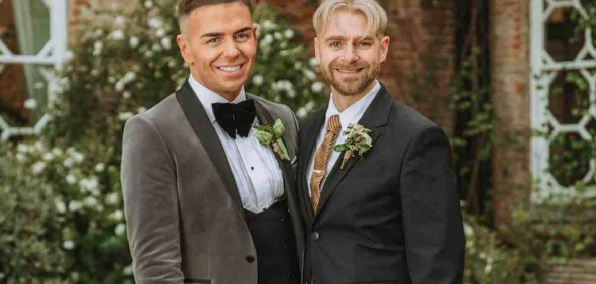A screenshot of Thomas and Adrian dressed in their wedding outfits from Married at First Sight UK. Thomas' grey suit looks to be made from velvet and he's wearing a formal white shirt and black bowtie and Adrian is dressed in a more conventional dark-looking suit with patterned tie.