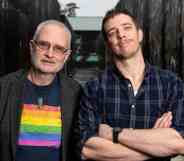 Eli Matthewson wears a dark, plaid patterned shirt as he crosses his arms in front of his chest. He is standing next to his dad who is wearing a shirt with a rainbow LGBTQ+ pride flag on it