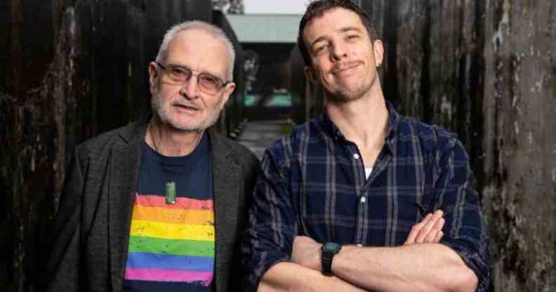 Eli Matthewson wears a dark, plaid patterned shirt as he crosses his arms in front of his chest. He is standing next to his dad who is wearing a shirt with a rainbow LGBTQ+ pride flag on it