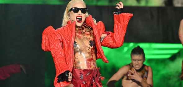 Lady Gaga performing on stage, wearing a thick red jacket with pointy shoulders over a see-through body