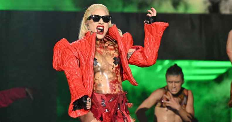 Lady Gaga performing on stage, wearing a thick red jacket with pointy shoulders over a see-through body