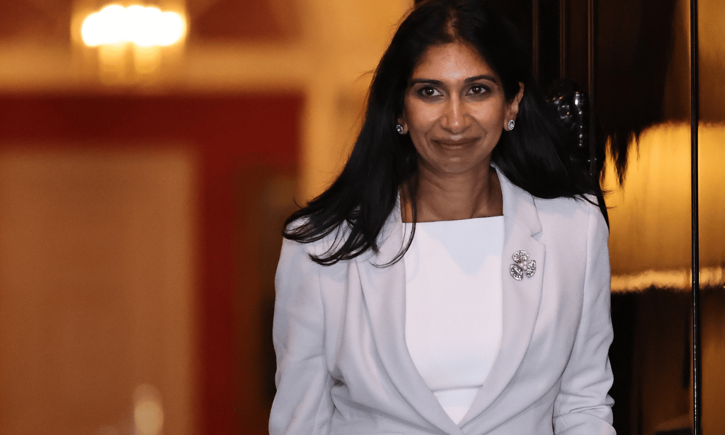 Suella Braverman wears a white and grey outfit as she walks out a doorway