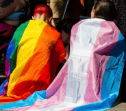 Two people sit side by side while wearing an LGBTQ+ and trans pride flag around their shoulders