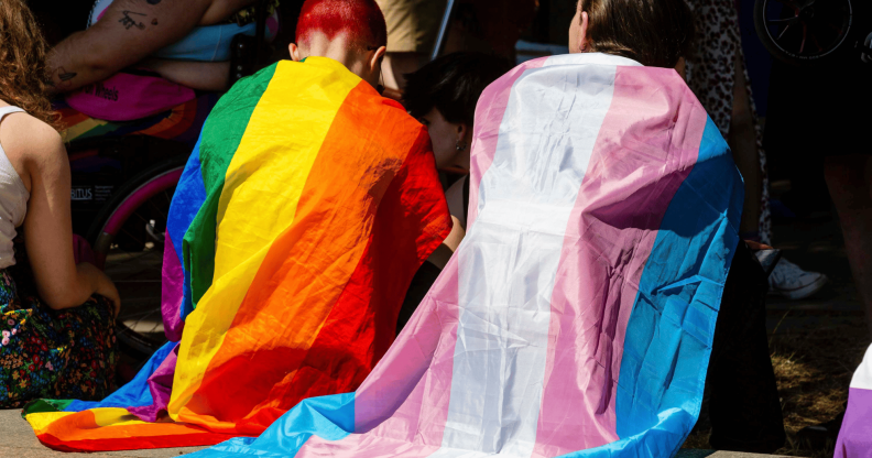 Two people sit side by side while wearing an LGBTQ+ and trans pride flag around their shoulders