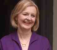 Liz Truss smiles as she stands outside a building wearing a purple outfit