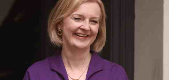 Liz Truss smiles as she stands outside a building wearing a purple outfit
