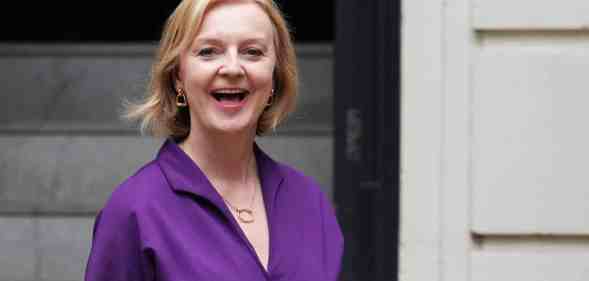 Liz Truss smiles as she stands outside a building wearing a purple outfit
