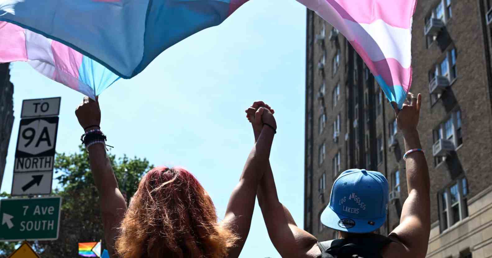 Two trans people hold hands as they march under a trans pride flag.