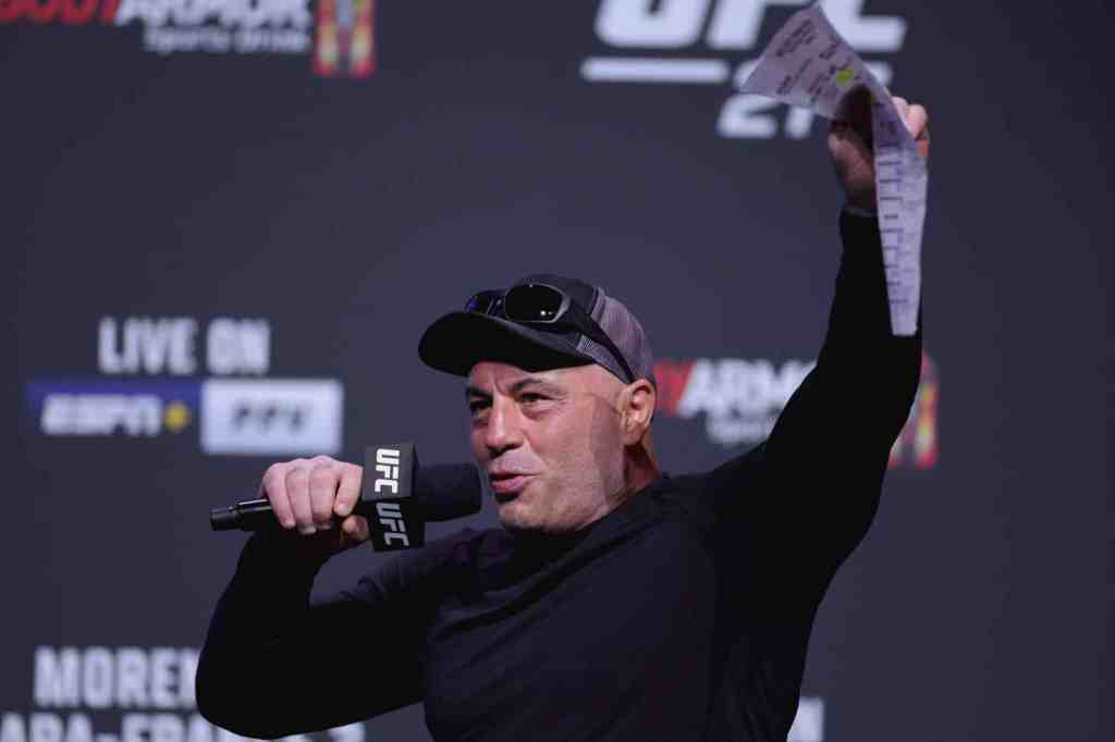 Joe Rogan raises a document in the air while speaking into a microphone excitedly.