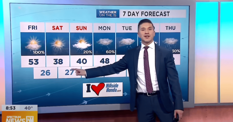A still of NY1 weatherman Erick Adame wearing a navy suit, white shirt and red tie giving one of his TV weather broadcasts
